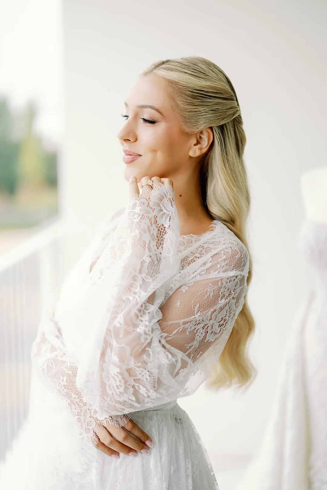 Noiva de cabelo loiro solto e maquilhagem natural, de olhos fechados junto a uma janela, com um vestido branco ao fundo